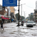 Rohtak waterlogging floods a city road after heavy monsoon rain, with vehicles and pedestrians moving through the water.
