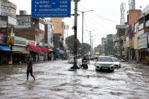 Rohtak waterlogging floods a city road after heavy monsoon rain, with vehicles and pedestrians moving through the water.